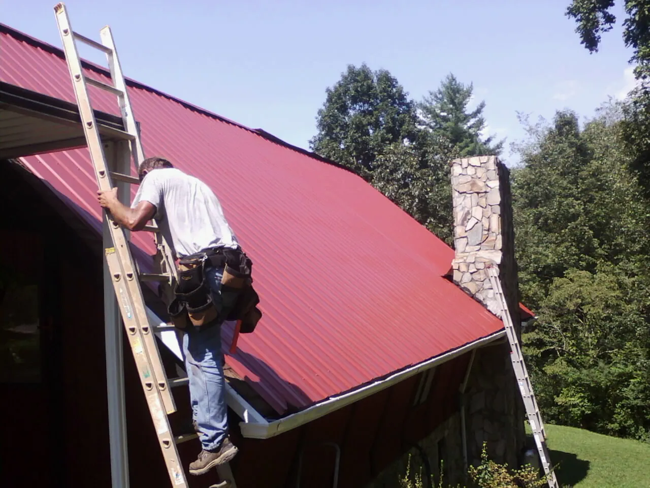 Roofer on a ladder beside a red metal roof and stone chimney — Soffit & Fascia Repair by Roofing Brothers in Kittanning