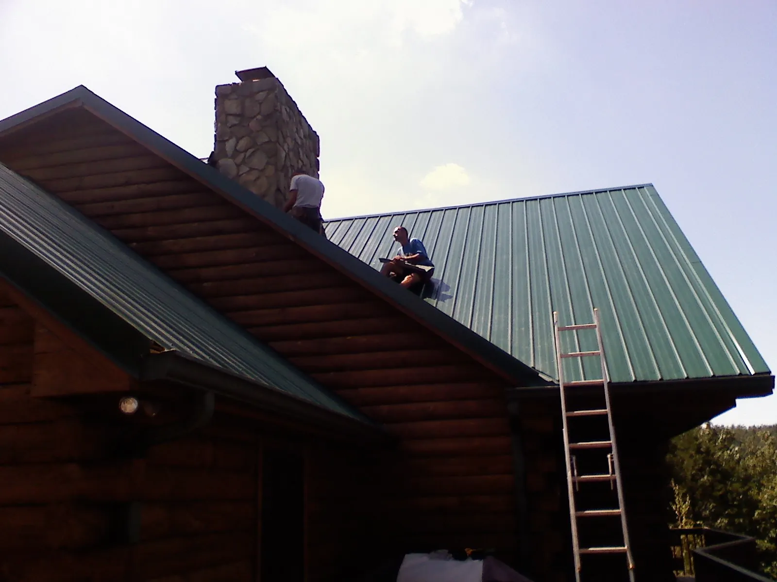 Roofers installing green metal panels beside a stone chimney on a log home in Kittanning